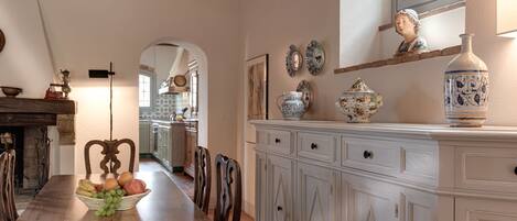 A dining area with a wooden table, chairs, and a fruit bowl. There is a white cabinet with decorative plates and a vase. A fireplace is visible in the background.