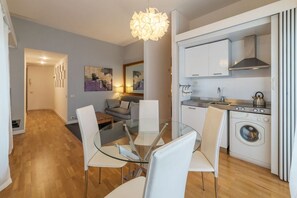 A modern kitchen with a built-in washing machine, a dining area with a glass table, and a living space with a sofa and artwork.