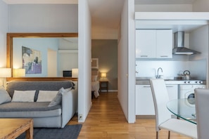 A compact living space with a sofa, a glass dining table, a kitchen area with cabinets and a stove, and a hallway leading to a bedroom.