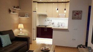 A compact living space with a sofa, a small table with a plate, a kitchen area with cabinets and a refrigerator, and a wall-mounted shelf with decorative items. - Charming apartment in the center of Malaga (Málaga)