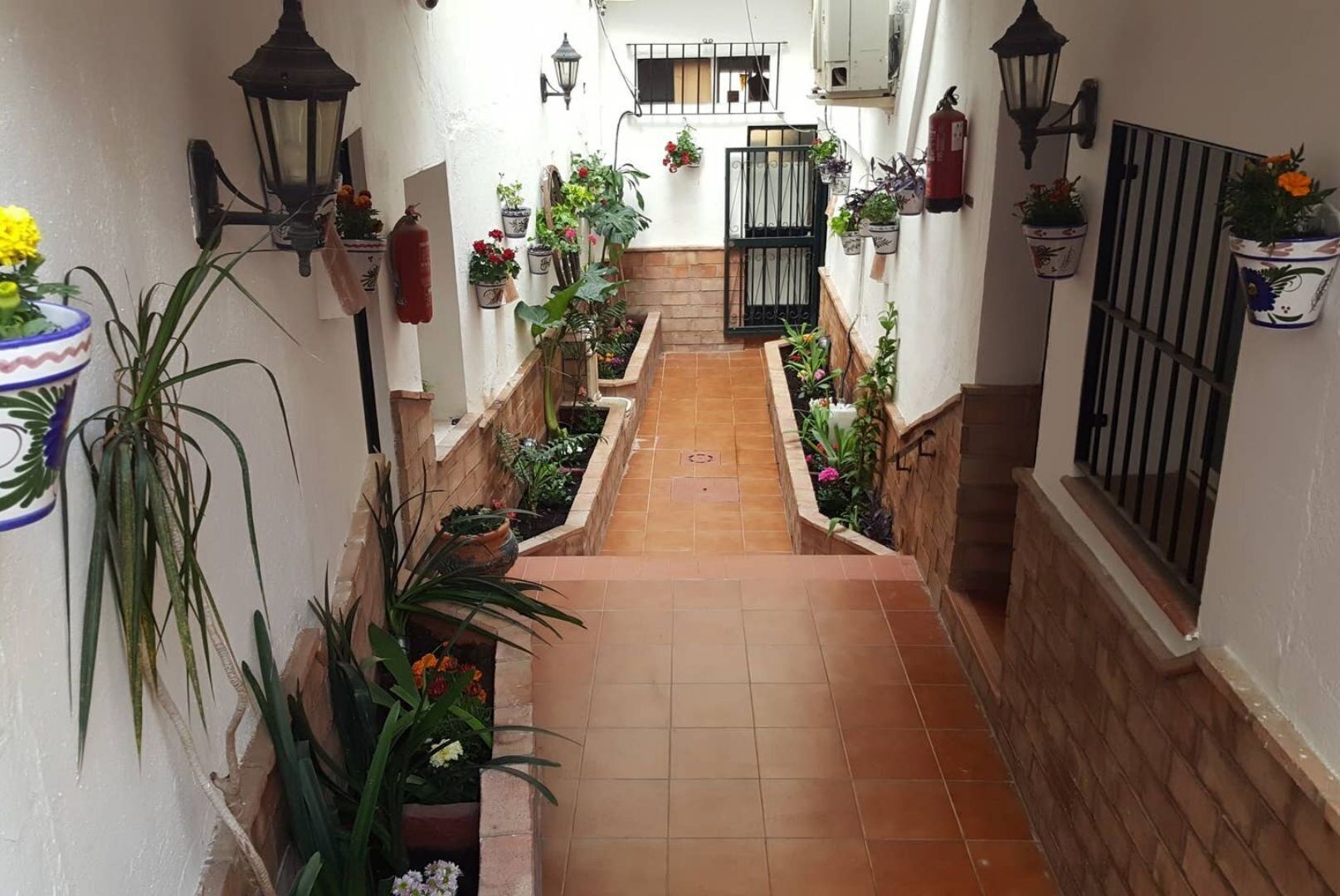 A narrow corridor with tiled flooring, potted plants on both sides, and a gate at the end.