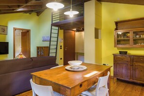 Um quarto acolhedor com uma mesa de jantar de madeira, cadeiras brancas, um sofá e uma área de cozinha com armários e uma luminária pendente.