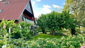 A house with a red-tiled roof, a garden with various plants and flowers, and a large tree. - Apartment East with balcony - Trabbi house (Wittmund)