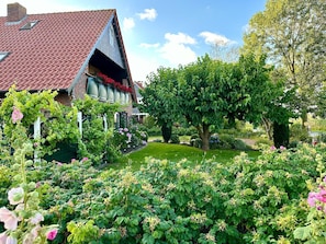 Une maison avec un toit recouvert de tuiles rouges, un jardin où poussent différentes plantes et fleurs, et un grand arbre.