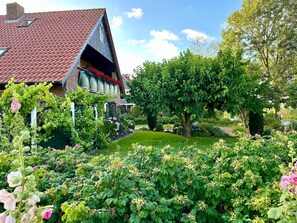 Une maison avec un toit recouvert de tuiles rouges, un jardin où poussent différentes plantes et fleurs, et un grand arbre.