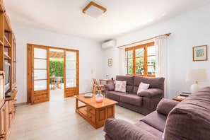 Amplio salón con sofá, una mesita de centro y televisor. Puertas de madera dan a un área exterior.