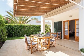 Eine Außenpatio mit einem Holztisch und -stühlen, einer Pergola und Blick auf einen Garten sowie einen Hauseingang.