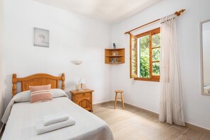A bedroom with a wooden bed, a nightstand, a small stool, a window with curtains, and a shelf with decorative items.