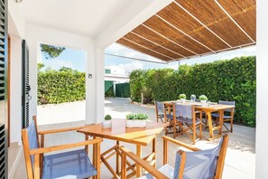 Eine Terrasse im Freien mit Holzmöbeln, einem für das Essen gedeckten Tisch und Blick auf einen Heckenstreifen und Gebäude im Hintergrund.