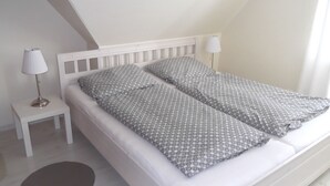 A neatly made twin bed with a patterned blanket, a bedside lamp, and a small bedside table in a simple room with white walls and a sloped ceiling. - Große Str. 2 Möwe, apt. 29 (Wyk auf Föhr)