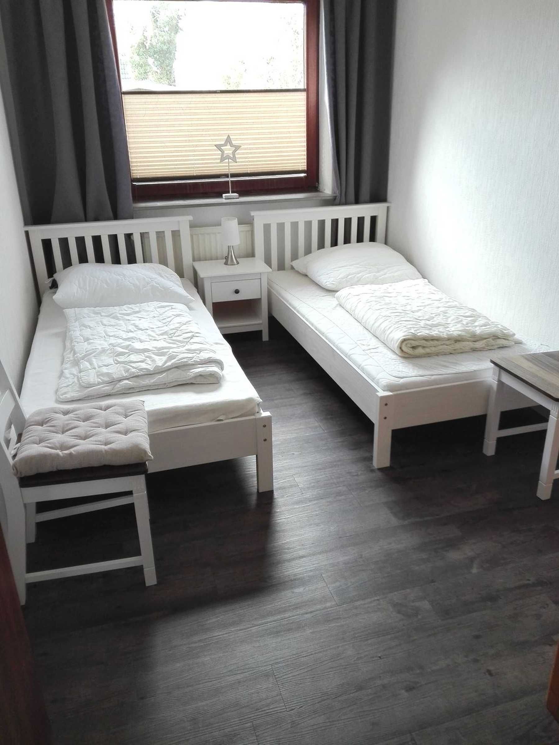 Quarto com duas camas de solteiro, uma pequena mesa de cabeceira com luminária, radiador e janela com cortinas.
