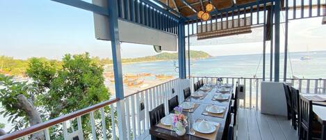 Un espacio exterior techado para comer con mesas dispuestas para dos, con vista a una playa y al mar.