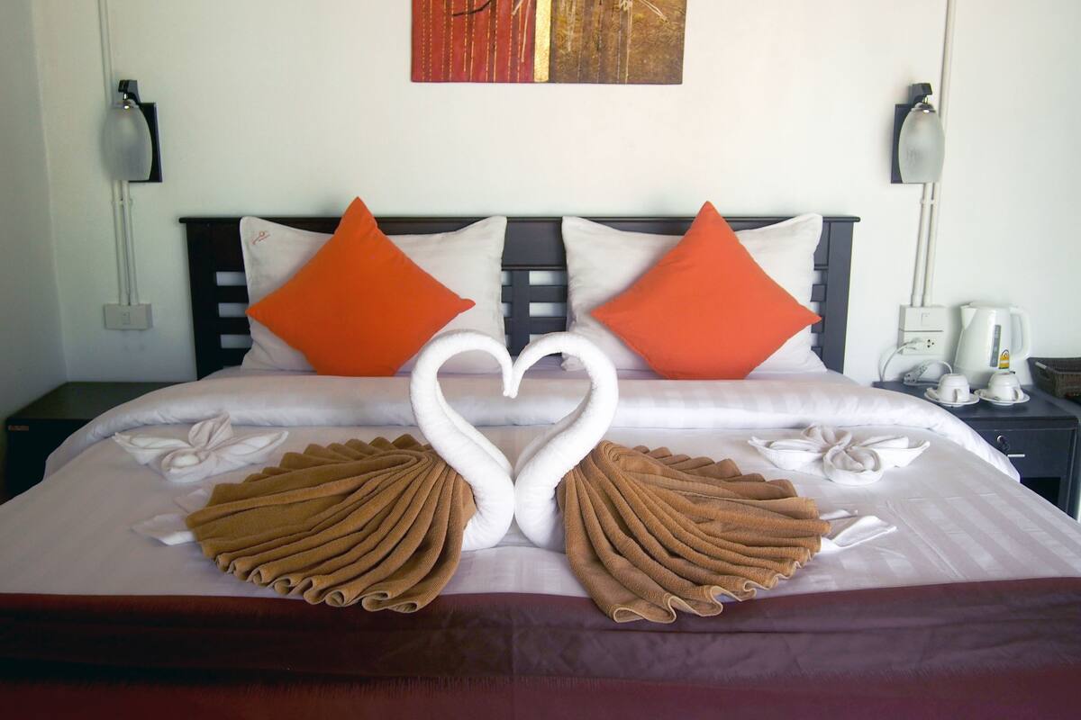 A neatly made bed with two swan-shaped towels, a painting on the wall, and a coffee maker on the nightstand.