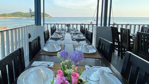 Un comedor con mesas dispuestas para una comida, con vistas a una playa con barcos y una vista despejada del mar.