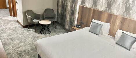 A hotel room with a large bed, two grey armchairs, a small round table, and a nightstand with a phone.