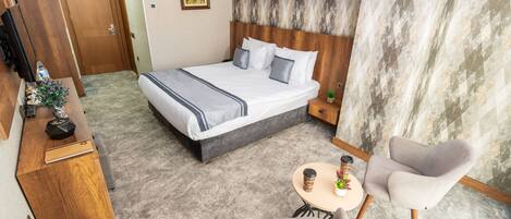 A hotel room with a bed, a desk, a chair, a small table with a cup, and a wall-mounted lamp.