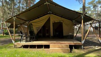 Een ruime tent met open zijkanten, een houten terras en stoelen, omringd door bomen.