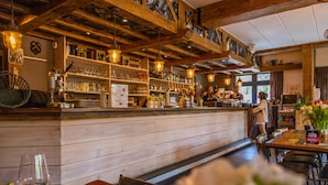 Un espace bar avec des poutres en bois, des étagères remplies de bouteilles et de verres, et un comptoir derrière lequel se trouve une personne.