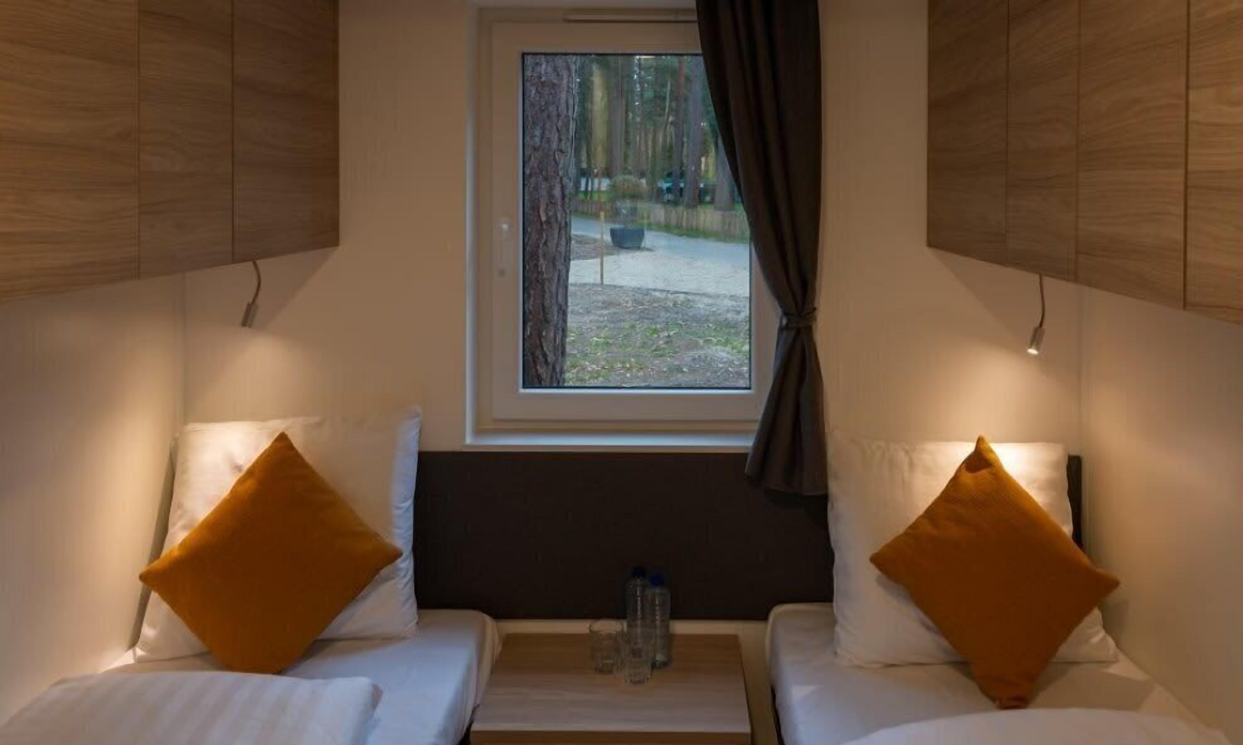 A small room with two beds, a window with a view of trees, and a table with a bottle and glasses.