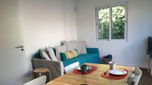 En hyggelig stue med spisebord til to, sofa med puder, et vindue med udsigt til grønne omgivelser og et fjernsyn på et stativ.