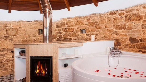 Una habitación de paredes de piedra con chimenea, una bañera grande con pétalos de rosa y una estructura de techo de madera.