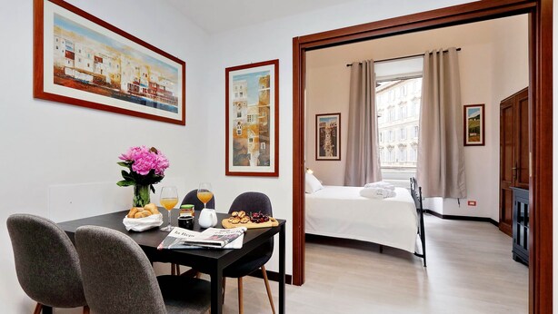 A hotel room with a bed, a dining area with a table, chairs, and a vase of flowers. There are framed pictures on the walls and a window with curtains. - Cavour Square Apartment (Rome)