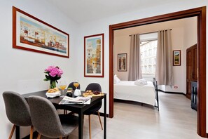 Habitación de hotel con cama, zona de comedor con mesa y sillas, y un jarrón de flores. Hay cuadros en las paredes y una ventana con cortinas.