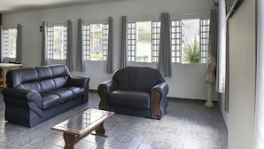 Amplia sala de estar con un sofá de cuero negro, una mesa de centro de madera y un sillón negro. Grandes ventanas con cortinas grises dejan entrar luz natural.