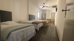 Habitación de hotel con dos camas individuales, una mesita pequeña, una silla, un ventilador de techo y un baño visible a través de una puerta abierta.