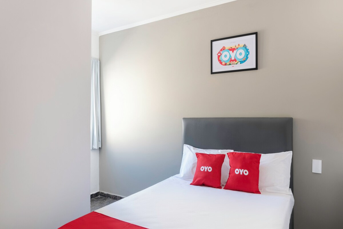 A hotel room with a bed, two pillows with 'OYO' branding, a framed picture on the wall, and a window with a curtain.