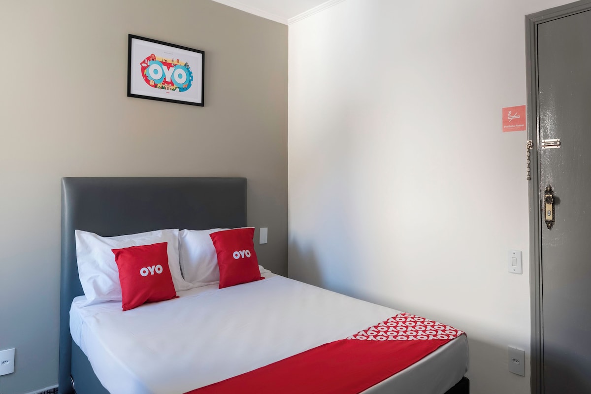 Habitación de hotel con una cama, almohadas con el logotipo del hotel, un cuadro enmarcado en la pared y una puerta con un cartel.