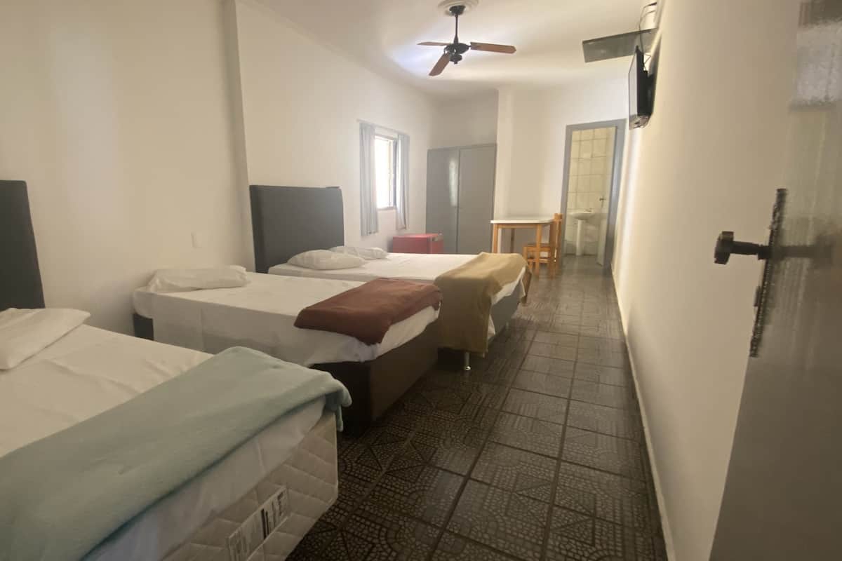 Ett hotellrum med tre sängar, en takfläkt, ett litet bord med en stol och ett badrum som syns genom en öppen dörr.