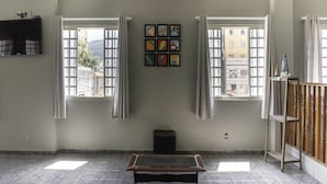 Una habitación con piso estampado, una mesa, una estantería con objetos y un ventanal con cortinas.