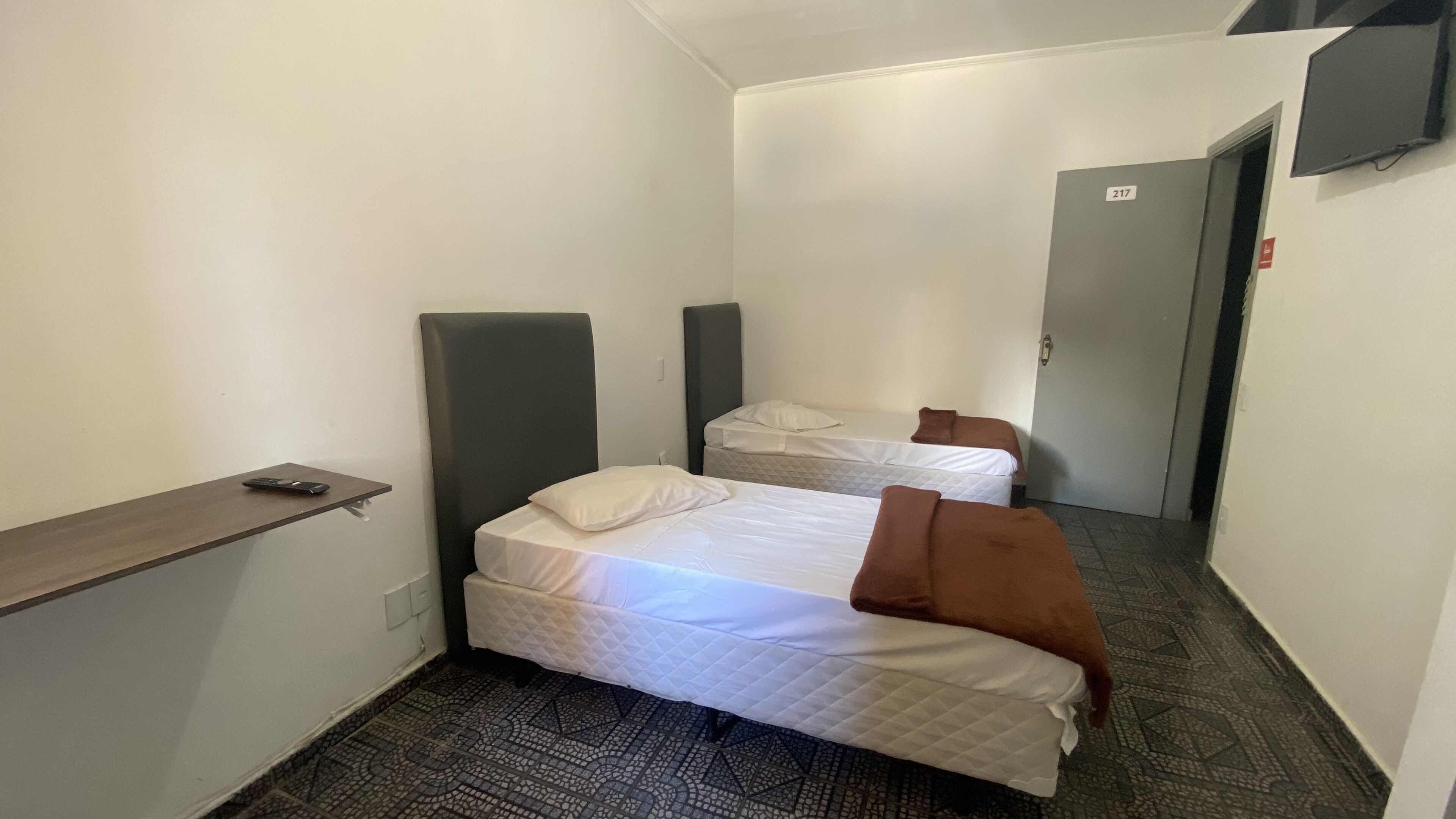 Una habitación de hotel pequeña y estrecha con dos camas, un escritorio y un televisor montado en la pared.