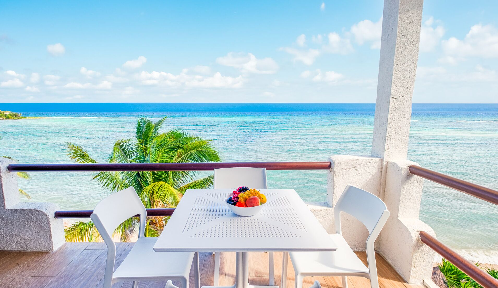 A balcony with a white table and chairs, a bowl of fruit, and a view of the ocean.