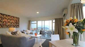 Ein modernes Wohnzimmer mit grauem Sofa, einem kleinen Tisch mit Blumenvase, einem Bild an der Wand und einem Balkon mit Blick.