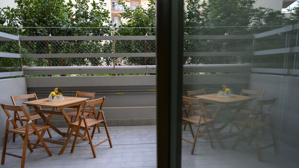 Un patio al aire libre con mesas y sillas de madera, una cerca de metal y vistas a una carretera y follaje.