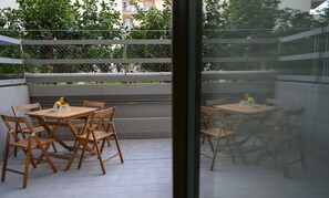 Een terras buiten met houten tafels en stoelen, een metalen hek en uitzicht op een weg en begroeiing.