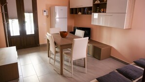 Una cocina compacta con refrigerador, una mesa de comedor con sillas, un televisor pequeño y un estante con objetos decorativos.