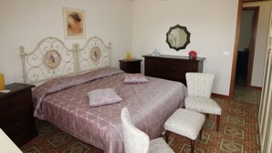 A bedroom with a bed, two bedside tables, a dresser, a chair, a mirror, and a hanging light fixture. - Casa Lungomare (Cefalù)