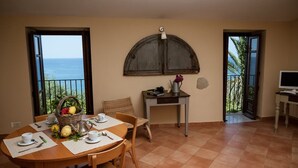 Zona pranzo con tavolo apparecchiato per due, un cesto di vimini con frutta, una caffettiera e vista sul mare attraverso porte aperte.