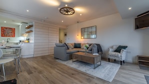 A modern living room with a grey sofa, a coffee table, and a patterned armchair. A kitchen area with white cabinets and a dining table is visible in the background. - Wheelwrights, Sustead, Norfolk (Sustead)