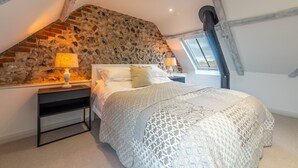 A bedroom with a sloped ceiling, exposed wooden beams, a stone wall, and a large window. - Wheelwrights, Sustead, Norfolk (Sustead)