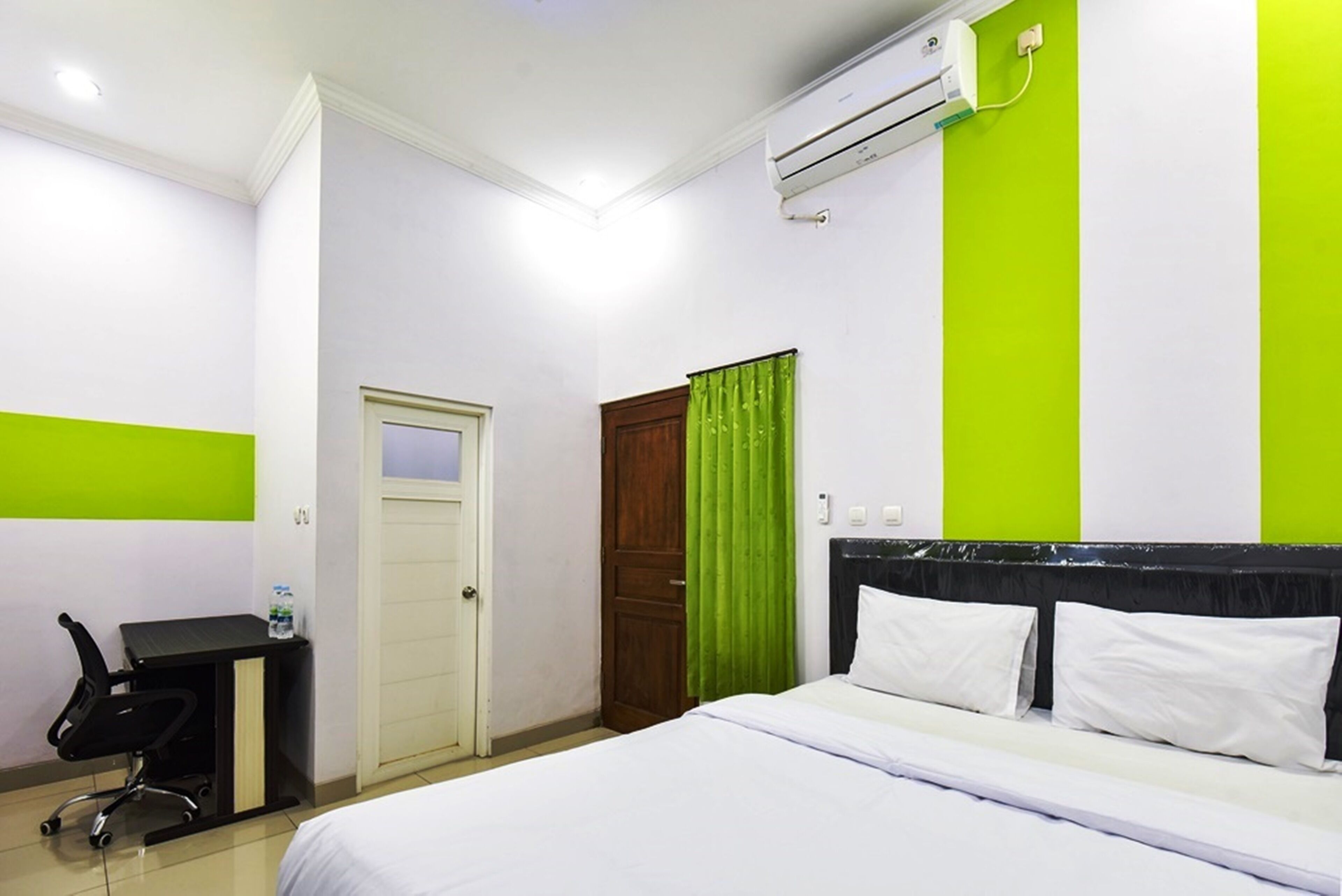 A hotel room with a bed, a desk with a chair, an air conditioning unit, and a green curtain.