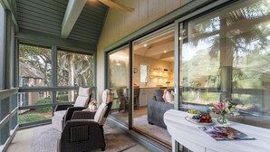 A spacious balcony with wicker furniture, a white table with a vase of flowers, and a view of the kitchen and dining area through large sliding glass doors.