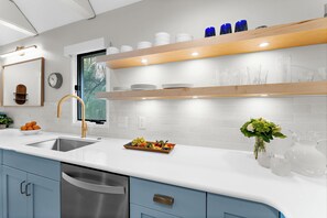 A modern kitchen with a white countertop, stainless steel sink, and blue cabinets. There are shelves above the counter holding glassware and a vase with flowers.