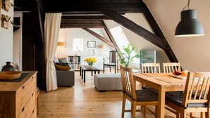 A cozy loft-style room with exposed wooden beams, a dining area with wooden furniture, and a view of a kitchen and living space through a large window. - Sunny mansard in the heart of Old Town (Centra rajons)