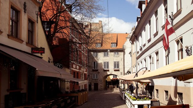 A cobblestone street with buildings on either side, a café with outdoor seating, and a flag on one of the buildings. - Sunny mansard in the heart of Old Town (Centra rajons)