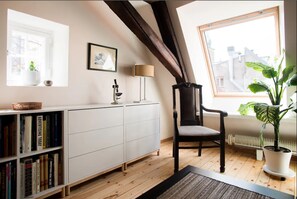 A room with a white bookshelf, a black chair, a lamp, and a large window with a view of buildings.
