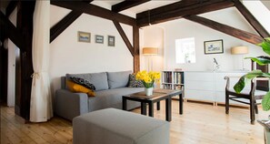 A cozy living room with a grey sofa, a coffee table with a vase of yellow flowers, a wooden floor, and exposed wooden beams.